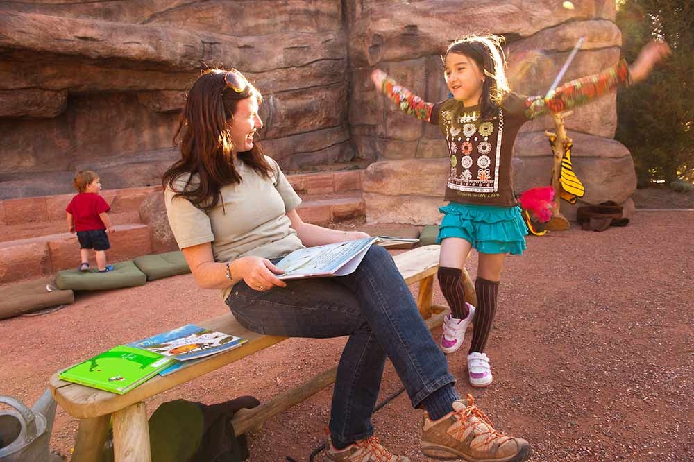 Smiling seated adult outside looking at child standing with her arms outreached. The adult has an open book in her lap. 