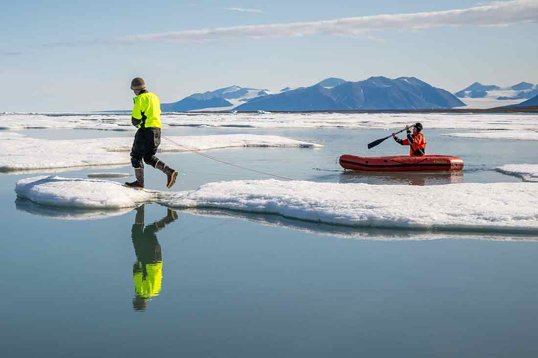 One person walking on snow and another paddling in an inflatable boat.