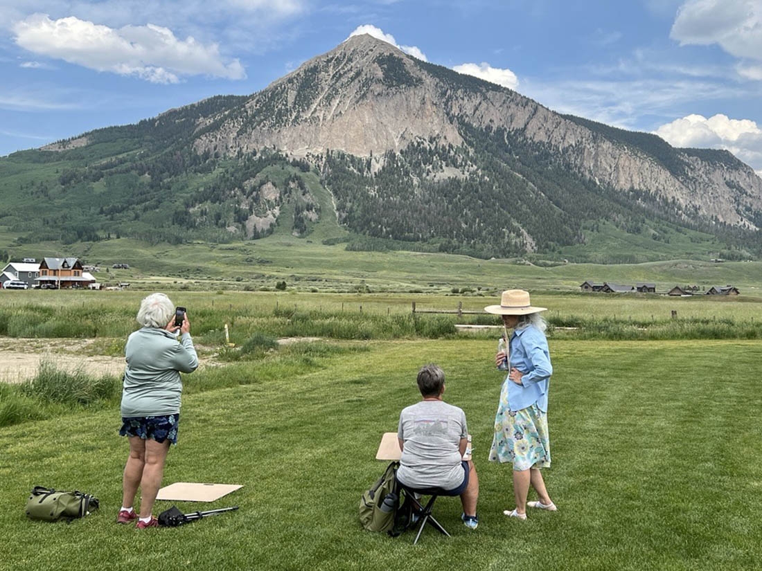 Three people observe a mountain in the distance and one sits and paints
