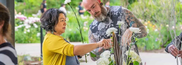 Woman arranging white flowers in a vase with a man instructing her.